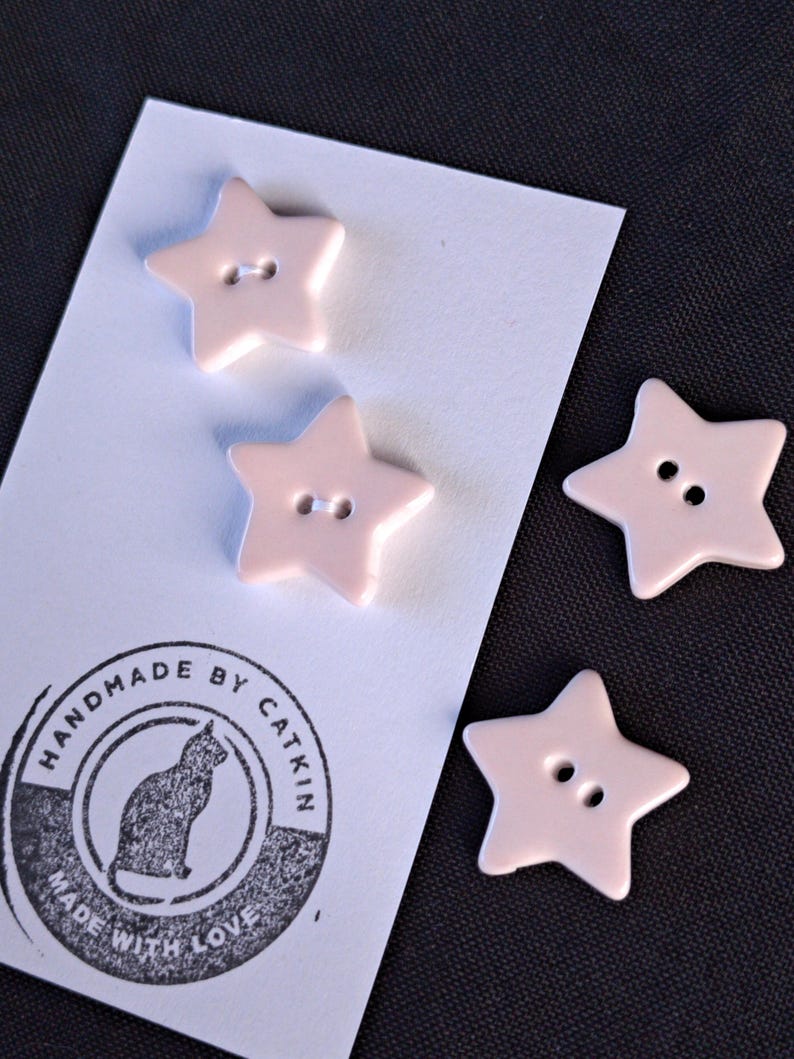 May include: Four light pink star-shaped buttons with two holes each. Two buttons are attached to a white rectangular card featuring a circular black stamp with a cat illustration and the text HANDMADE BY CATKIN MADE WITH LOVE.