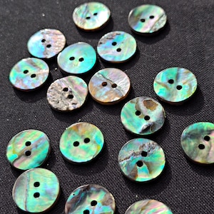 May include: A collection of round, two-hole buttons crafted from iridescent abalone shell. Each button displays a unique, swirling pattern of green, blue, and purple hues with a polished, reflective surface, showcasing the natural texture of the shell.