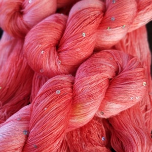 May include: Close-up of a soft, pink yarn with iridescent sequins woven throughout. The yarn is twisted and piled together, showing the texture and color variations.
