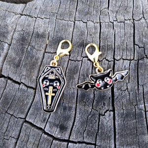 May include: Two gold-tone charms, one shaped like a coffin with a cross and the other shaped like a bat with red eyes. Both charms have black enamel and are attached to gold-tone lobster clasps.