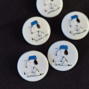 May include: Four circular white buttons featuring a graphic of the cartoon character Snoopy wearing a blue baseball cap. Each button shows the character in a different pose, including sitting and standing, with green grass details at the base.