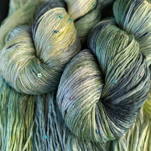 Puede incluir: Una madeja de hilo teñido a mano con una paleta de colores variados en verde salvia, verde bosque y amarillo pálido. El hilo está retorcido en un paquete e incluye pequeñas lentejuelas iridiscentes incrustadas que captan la luz en las fibras.