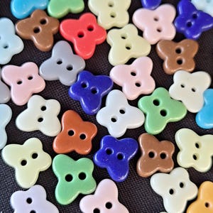 May include: A collection of butterfly-shaped buttons in various colours including blue, green, pink, brown, red, and white. Each button features two holes in the centre for attachment, with a smooth finish and distinct wing-like shapes.