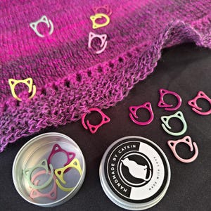 May include: A collection of colorful cat-shaped stitch markers in a small tin, with a matching knitted shawl in the background. The markers are in shades of pink, yellow, and green. The tin lid reads "HANDMADE BY CATKIN MADE WITH LOVE."