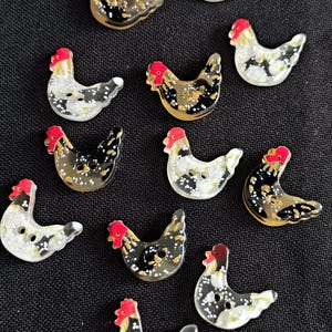 May include: A selection of chicken-shaped buttons. Each button has a red comb, a speckled black and white body, and a buttonhole. The buttons are displayed on a black background.