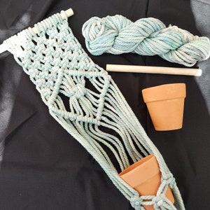 May include: A macrame plant hanger made with light blue rope, featuring a geometric design. The hanger is hanging from a wooden dowel and has two terracotta pots.