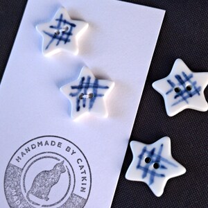 May include: Four star-shaped buttons with a white base and blue grid pattern. Each button features two holes in the center. The buttons are attached to a white display card featuring a circular stamp with a cat silhouette and the text HANDMADE BY CATKIN MADE WITH LOVE.
