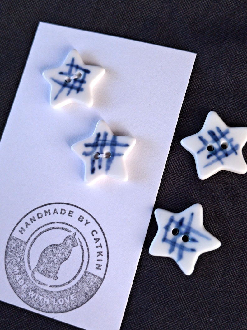 May include: Four star-shaped buttons with a white base and blue grid pattern. Each button features two holes in the center. The buttons are attached to a white display card featuring a circular stamp with a cat silhouette and the text HANDMADE BY CATKIN MADE WITH LOVE.