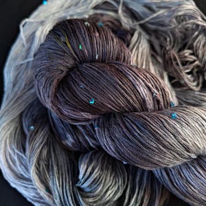 May include: A skein of hand-dyed yarn featuring a gradient transition from deep charcoal grey to light silver. The yarn is speckled with small, vibrant turquoise blue metallic flecks throughout the twisted fibres, creating a textured appearance.