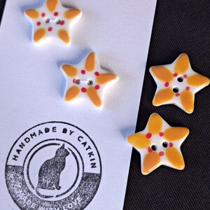 May include: Four star-shaped buttons with a white base, yellow petal-like accents, and small red dots around the center holes. The buttons are attached to a white card featuring a circular black stamp that reads HANDMADE BY CATKIN MADE WITH LOVE.