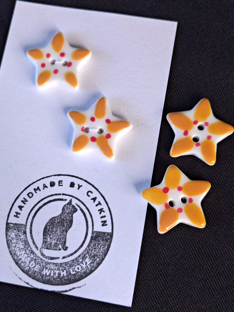 May include: Four star-shaped buttons with a white base, yellow petal-like accents, and small red dots around the center holes. The buttons are attached to a white card featuring a circular black stamp that reads HANDMADE BY CATKIN MADE WITH LOVE.
