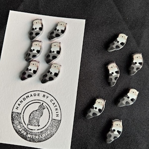 Könnte beinhalten: Handgefertigte Otter-Anstecknadeln mit schwarzen, weißen und grauen Mustern. Sie sind auf einer weißen Karte mit einem runden Stempel mit der Aufschrift "HANDMADE BY CATKIN" und "MADE WITH LOVE" angeordnet.