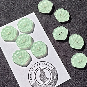 May include: A set of light green, translucent, shell-shaped buttons with embedded glitter. Five buttons are attached to a white card featuring a circular logo with a cat silhouette and the text "HANDMADE BY CATKIN MADE WITH LOVE". Four additional buttons are loose.