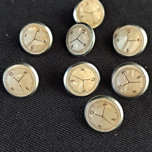 May include: A set of seven circular buttons featuring a cream-coloured mother-of-pearl inlay with a printed clock face design. Each button has a metallic silver-toned rim and displays black hour markers and hands indicating a time on the dial.