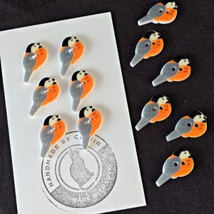 May include: A collection of bird-shaped buttons featuring a black head, grey wing, and orange breast. Six buttons are attached to a white rectangular card with a circular stamp reading HANDMADE BY CATKIN MADE WITH LOVE, and four loose buttons are nearby.
