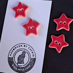 May include: Four red star-shaped buttons with two holes each. Two buttons are attached to a white rectangular card featuring a black circular stamp with a cat illustration and the text HANDMADE BY CATKIN MADE WITH LOVE. Two buttons are loose.