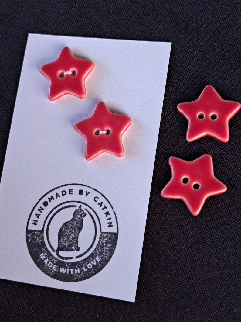 May include: Four red star-shaped buttons with two holes each. Two buttons are attached to a white rectangular card featuring a black circular stamp with a cat illustration and the text HANDMADE BY CATKIN MADE WITH LOVE. Two buttons are loose.