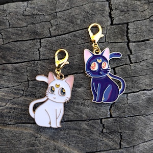 May include: Two enamel cat charms with gold-colored clasps. One is white with pink accents, the other is dark blue with pink accents. Both have a crescent moon on their foreheads and large, expressive eyes. The charms are set against a dark, textured background.