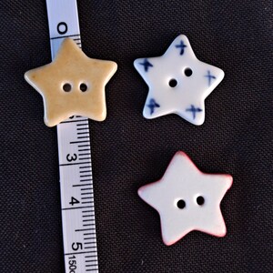 May include: Three star-shaped buttons with two holes each. One button is tan, one is white with blue cross patterns, and one is white with a red edge. A white measuring tape is positioned next to the tan button, showing a width of 1.25 inches.
