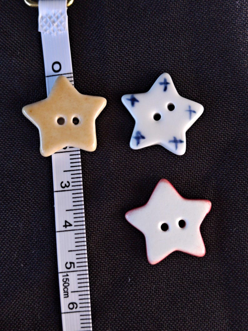 May include: Three star-shaped buttons with two holes each. One button is tan, one is white with blue cross patterns, and one is white with a red edge. A white measuring tape is positioned next to the tan button, showing a width of 1.25 inches.
