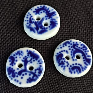 May include: Three white ceramic buttons with blue floral patterns and two holes each. The buttons are round and have a glossy finish. The blue design is intricate and detailed, set against a white background.