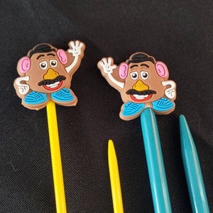May include: Two plastic toy Mr. Potato Head figures with blue and white clothing, attached to yellow and blue knitting needles.