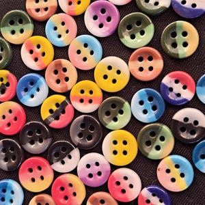 May include: A collection of round, four-hole buttons scattered across a dark surface. Each button features a distinct two-tone colour design, showcasing various combinations of yellow, pink, blue, green, purple, black, and white in a smooth finish.