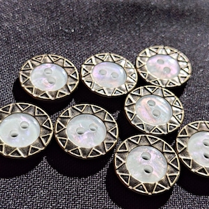 May include: A collection of seven round buttons, each featuring a central iridescent white inlay surrounded by a metallic bronze-toned rim with a repeating triangular geometric pattern. Every button has two small holes in the centre for attachment.