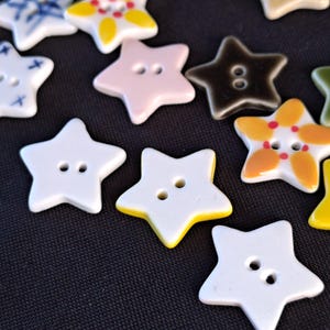 May include: A collection of star-shaped buttons in various colors and patterns, including solid white, yellow, pink, and dark brown, as well as white buttons with blue or yellow and red floral-like designs, each featuring two central holes.