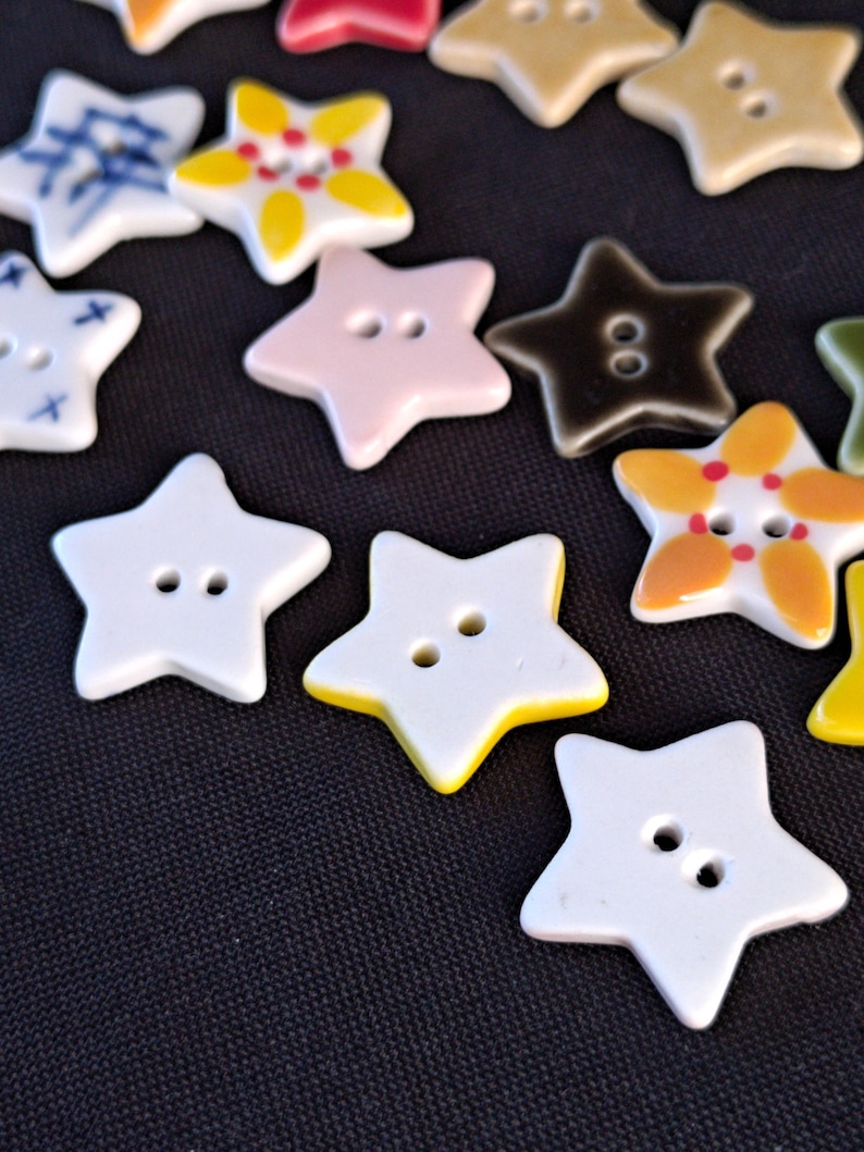 May include: A collection of star-shaped buttons in various colors and patterns, including solid white, yellow, pink, and dark brown, as well as white buttons with blue or yellow and red floral-like designs, each featuring two central holes.