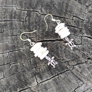 Peut inclure: Boucles d'oreilles en coquillages blancs avec un pendentif en forme de tortue argentée. Les boucles d'oreilles sont suspendues à une surface en bois.