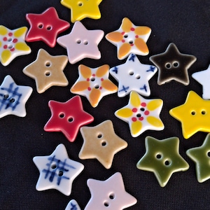 May include: A collection of star-shaped buttons in various colors and patterns, including solid yellow, red, pink, olive green, and brown, alongside white buttons featuring blue plaid, floral, or cross-stitch designs, each with two central holes.