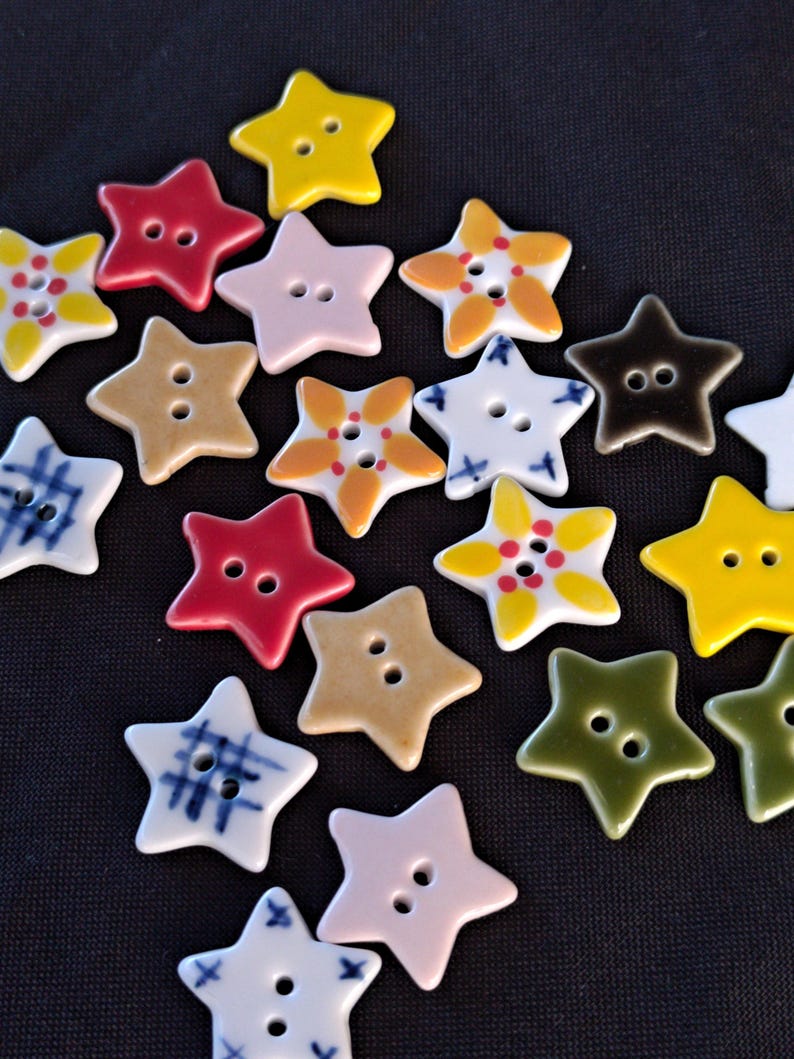 May include: A collection of star-shaped buttons in various colors and patterns, including solid yellow, red, pink, olive green, and brown, alongside white buttons featuring blue plaid, floral, or cross-stitch designs, each with two central holes.