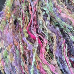 May include: Close-up of colourful yarn with a textured, looped appearance. The yarn features a blend of vibrant colours, including pink, green, purple, and yellow. The strands are tightly twisted and create a soft, fuzzy texture, ideal for fibre art projects.