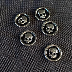 May include: Five circular buttons featuring a raised silver-toned skull emblem centred on a smooth black background. Each button is framed by a polished silver-toned metal rim, creating a distinct contrast between the dark centre and the metallic edge.