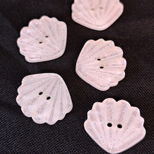 May include: Five light pink, shell-shaped buttons with two holes in the center. Each button features a translucent finish with embedded fine glitter particles that catch the light, creating a subtle shimmering effect across the ridged surface.