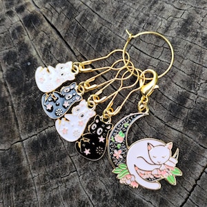 May include: A set of six gold cat stitch markers with black and white enamel details. The markers feature a crescent moon with a sleeping cat and a variety of cat designs.