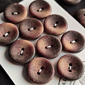 May include: A collection of round, two-hole buttons featuring a mottled, gradient finish that transitions from dark brown and black edges to a lighter, speckled tan centre. Each button has a concave surface and is attached to a white display card.