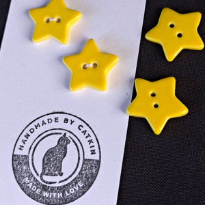 May include: Four bright yellow star-shaped buttons with two holes each. Two buttons are attached to a white rectangular card featuring a circular black ink stamp with a cat silhouette and the text HANDMADE BY CATKIN MADE WITH LOVE.