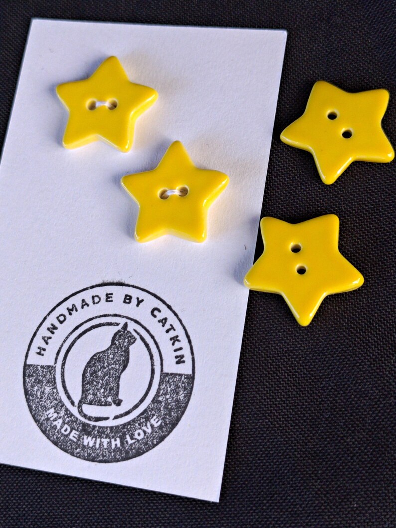May include: Four bright yellow star-shaped buttons with two holes each. Two buttons are attached to a white rectangular card featuring a circular black ink stamp with a cat silhouette and the text HANDMADE BY CATKIN MADE WITH LOVE.