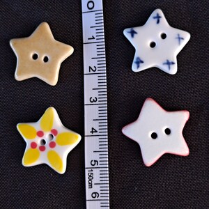 May include: Four star-shaped buttons with two holes each, arranged next to a white measuring tape showing centimeters. One button is tan, one is white with blue crosses, one is white with yellow petals and red dots, and one is white with a red edge.
