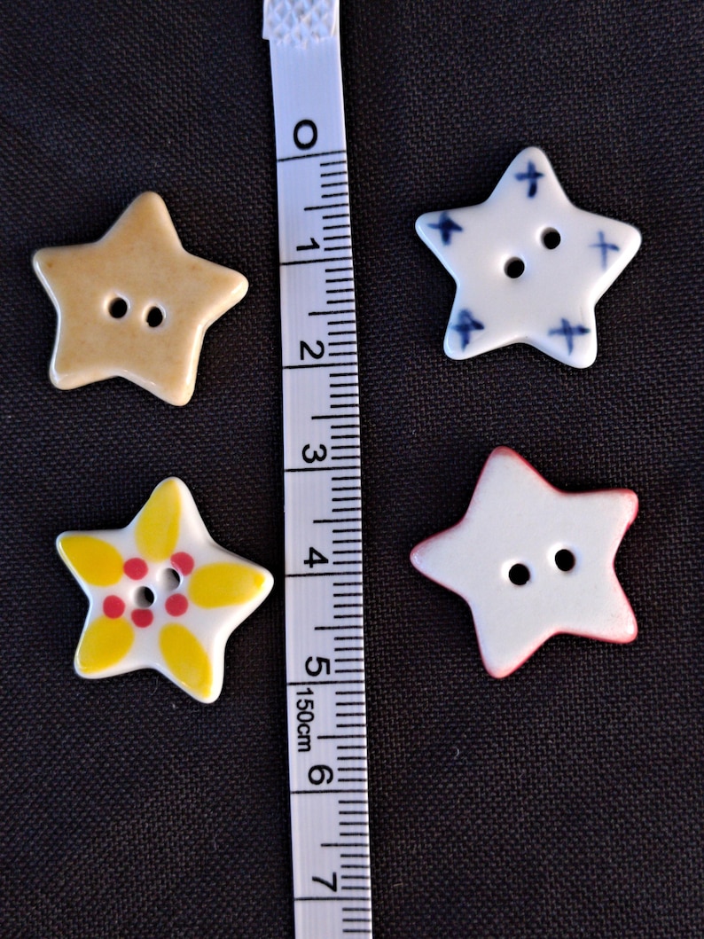 May include: Four star-shaped buttons with two holes each, arranged next to a white measuring tape showing centimeters. One button is tan, one is white with blue crosses, one is white with yellow petals and red dots, and one is white with a red edge.