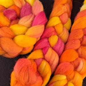 Könnte beinhalten: Nahaufnahme von leuchtenden, geflochtenen Fasern in Gelb-, Orange- und Rosatönen. Die Faser ist dick und flauschig, mit einer weichen Textur. Die Zöpfe sind kreisförmig angeordnet und erzeugen ein ansprechendes Design. Der Hintergrund ist dunkel.