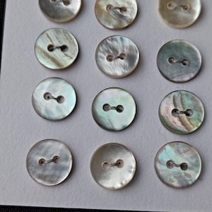 May include: A collection of twelve iridescent, round buttons with two holes each. The buttons have a pearlescent sheen, with colors ranging from white to pale green and gold. They are arranged in three rows of four on a light-colored card.