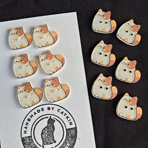 May include: A collection of cat-shaped buttons featuring a cream, orange, and brown color palette. Six buttons are attached to a white display card with the text "HANDMADE BY CATKIN MADE WITH LOVE" and a cat silhouette, while five buttons are loose.