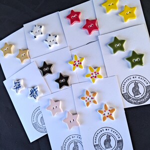 May include: A collection of star-shaped buttons in various colors and patterns, including solid red, yellow, olive green, and light pink, alongside white buttons with blue cross-hatch or floral designs, all mounted on white cards with a cat logo.