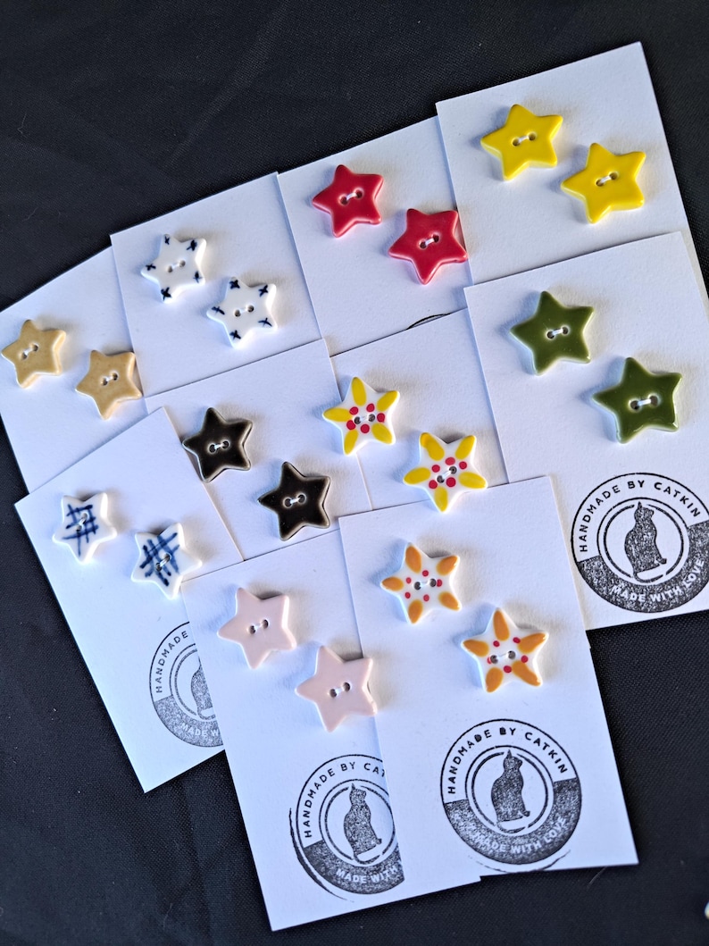 May include: A collection of star-shaped buttons in various colors and patterns, including solid red, yellow, olive green, and light pink, alongside white buttons with blue cross-hatch or floral designs, all mounted on white cards with a cat logo.