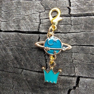 May include: A gold charm with a blue planet and a crown. The planet has a ring and a crescent moon. The crown has five points and is decorated with stars.