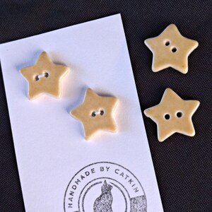 May include: Three star-shaped buttons in a light tan color, each featuring two central holes. Two buttons are attached to a white rectangular card with a circular stamp that reads "HANDMADE BY CATKIN MADE WITH LOVE" and a cat silhouette.