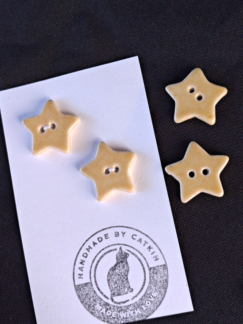 May include: Three star-shaped buttons in a light tan color, each featuring two central holes. Two buttons are attached to a white rectangular card with a circular stamp that reads "HANDMADE BY CATKIN MADE WITH LOVE" and a cat silhouette.